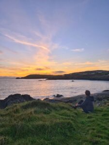Owenahincha Beach in West Cork, Irland im letzten Licht der untergehenden Sonne, eine Person sitzt am Ufer und beobachtet den Sonnenuntergang.